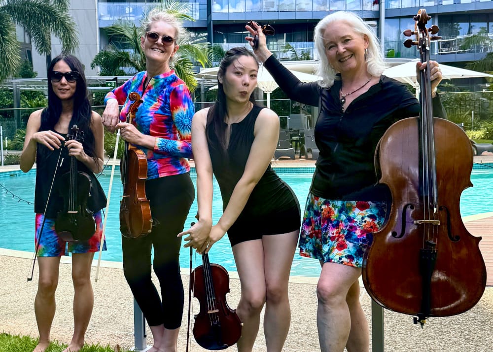 A group of four smiling women in OceanRoad swimwear posing poolside with violins and a cello during a creative brand collaboration.