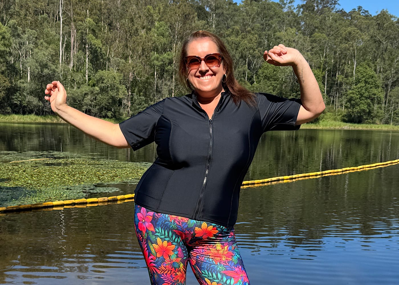 Laura from Timber Heart Creations standing by a lake with her arms raised in a celebratory pose. She is smiling and wearing a black short sleeve zip-up rashie and vibrant floral print swim leggings from OceanRoad Swimwear.