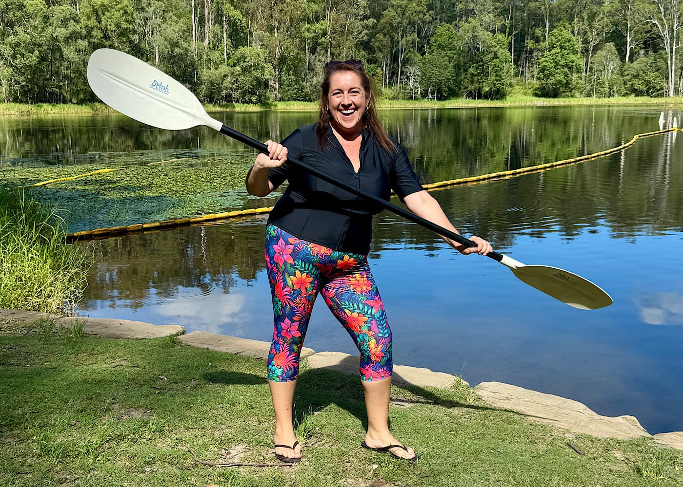  Laura Vincent standing lakeside holding a kayak paddle, smiling at the camera in OceanRoad swimwear.
